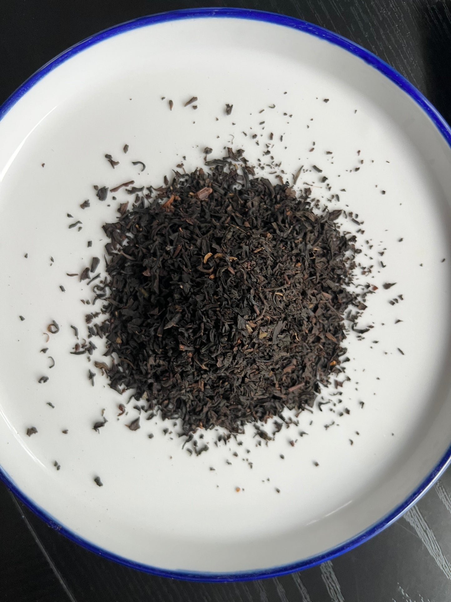 A pile of dark, finely ground loose-leaf tea or coffee beans sits in the center of a white ceramic plate with a blue rim, set against a dark wood background.
