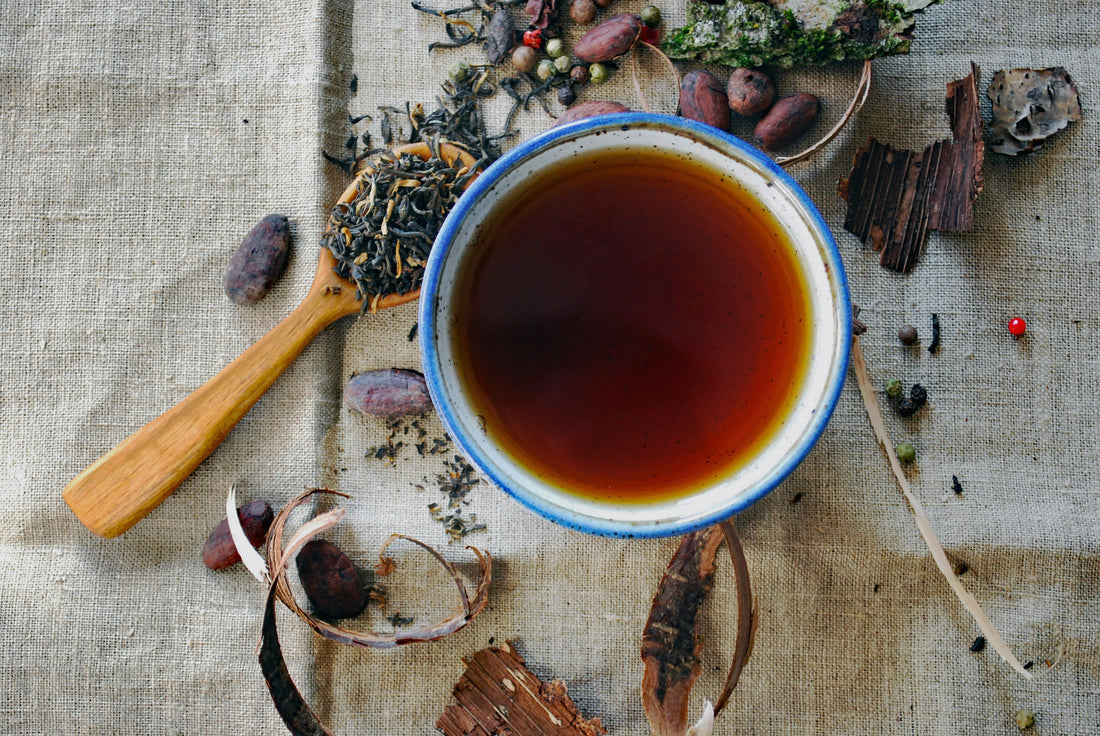 Overhead view of brewed herbal tea in blue-rimmed cup surrounded by loose tea leaves, herbs, spices and botanicals on linen - showing flavor profiles and ingredient variety for tea matching