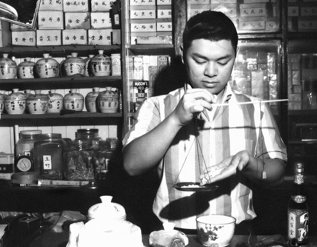 Traditional tea ceremony in black and white showing person carefully preparing tea in apothecary setting with jars and traditional tools - representing global tea culture and mindful rituals