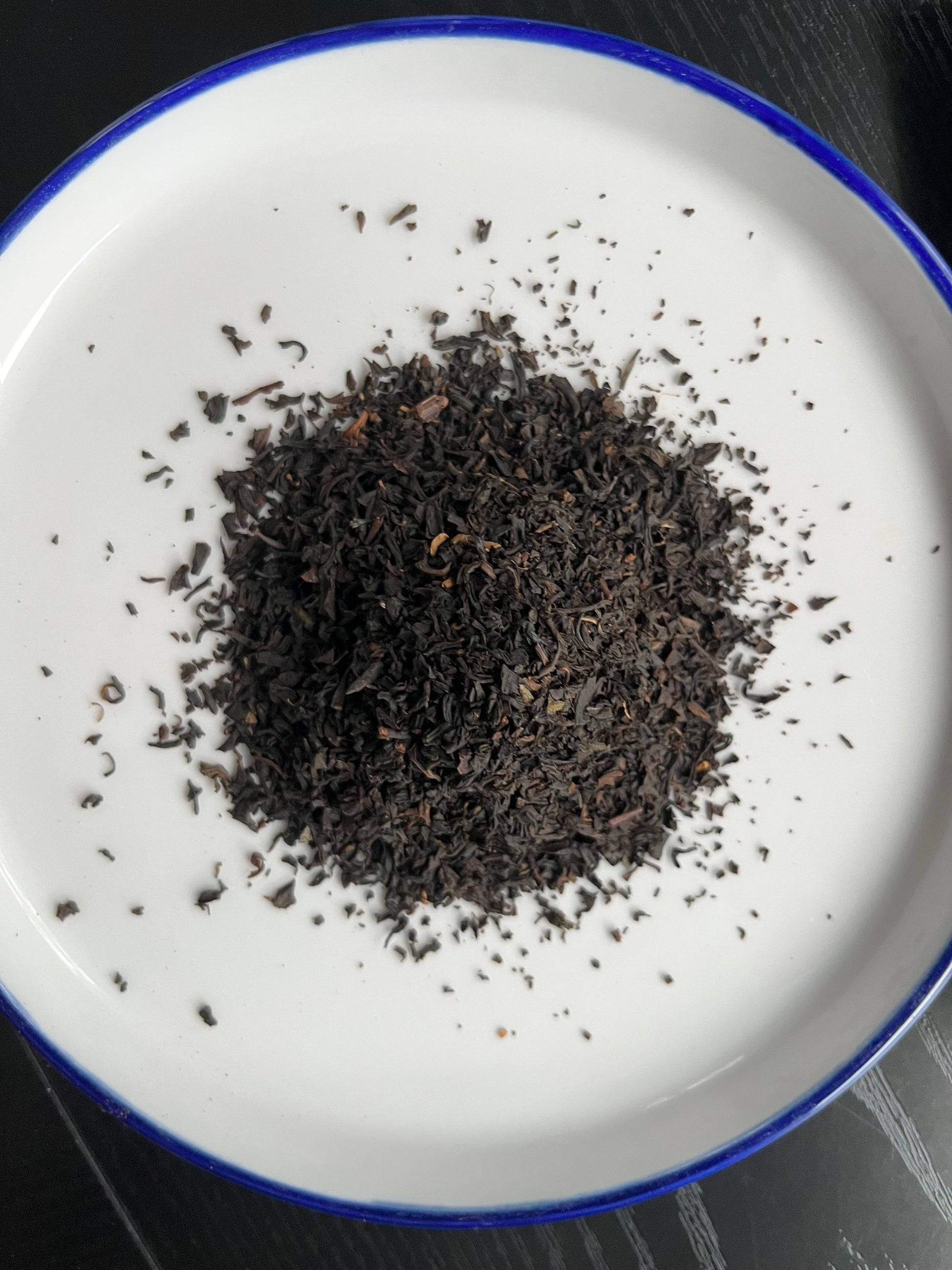 A pile of dark, finely ground loose-leaf tea or coffee beans sits in the center of a white ceramic plate with a blue rim, set against a dark wood background.