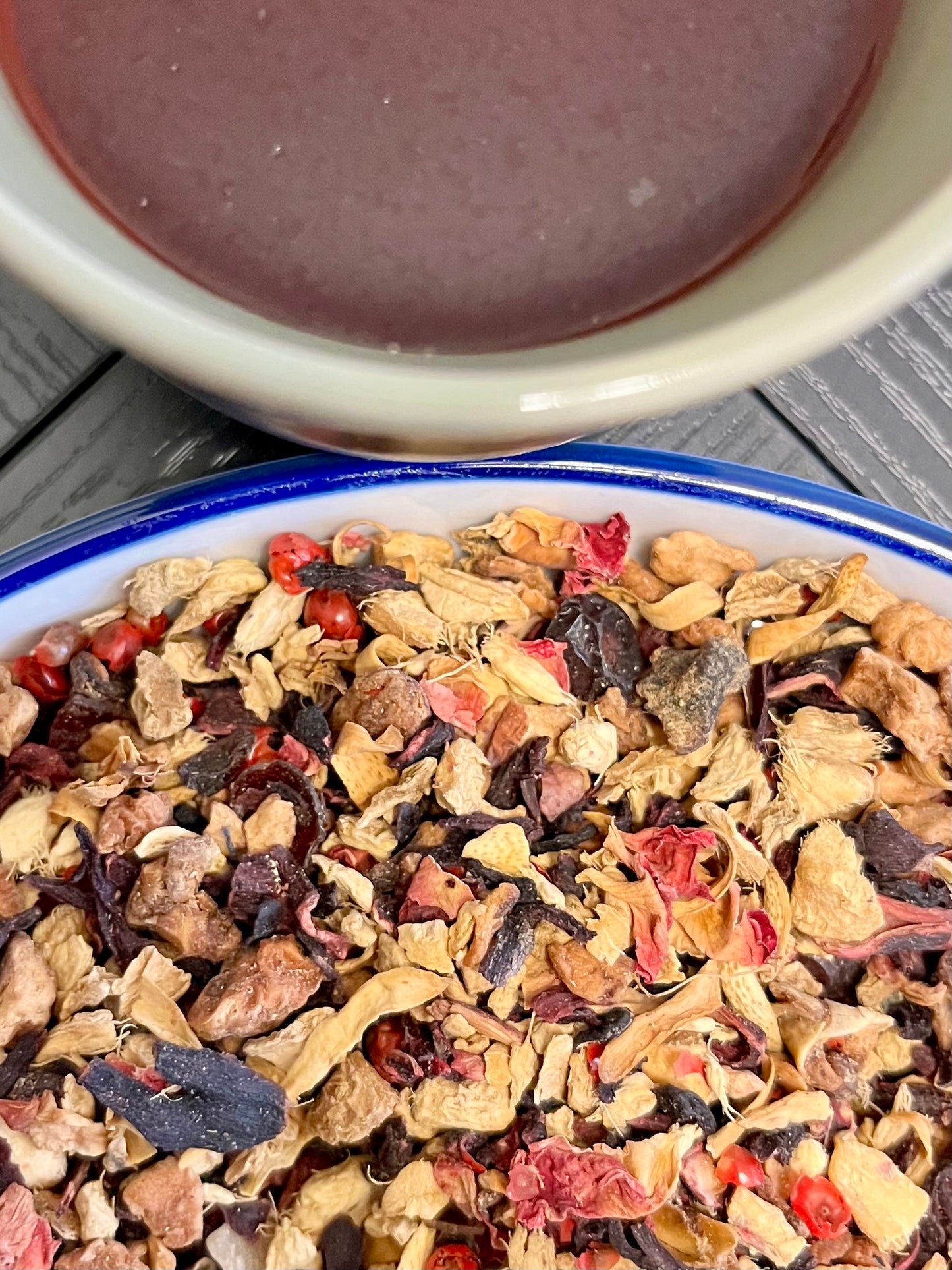 Tea leaves with visible herbs and fruits in a bowl, next to a cup of tea.