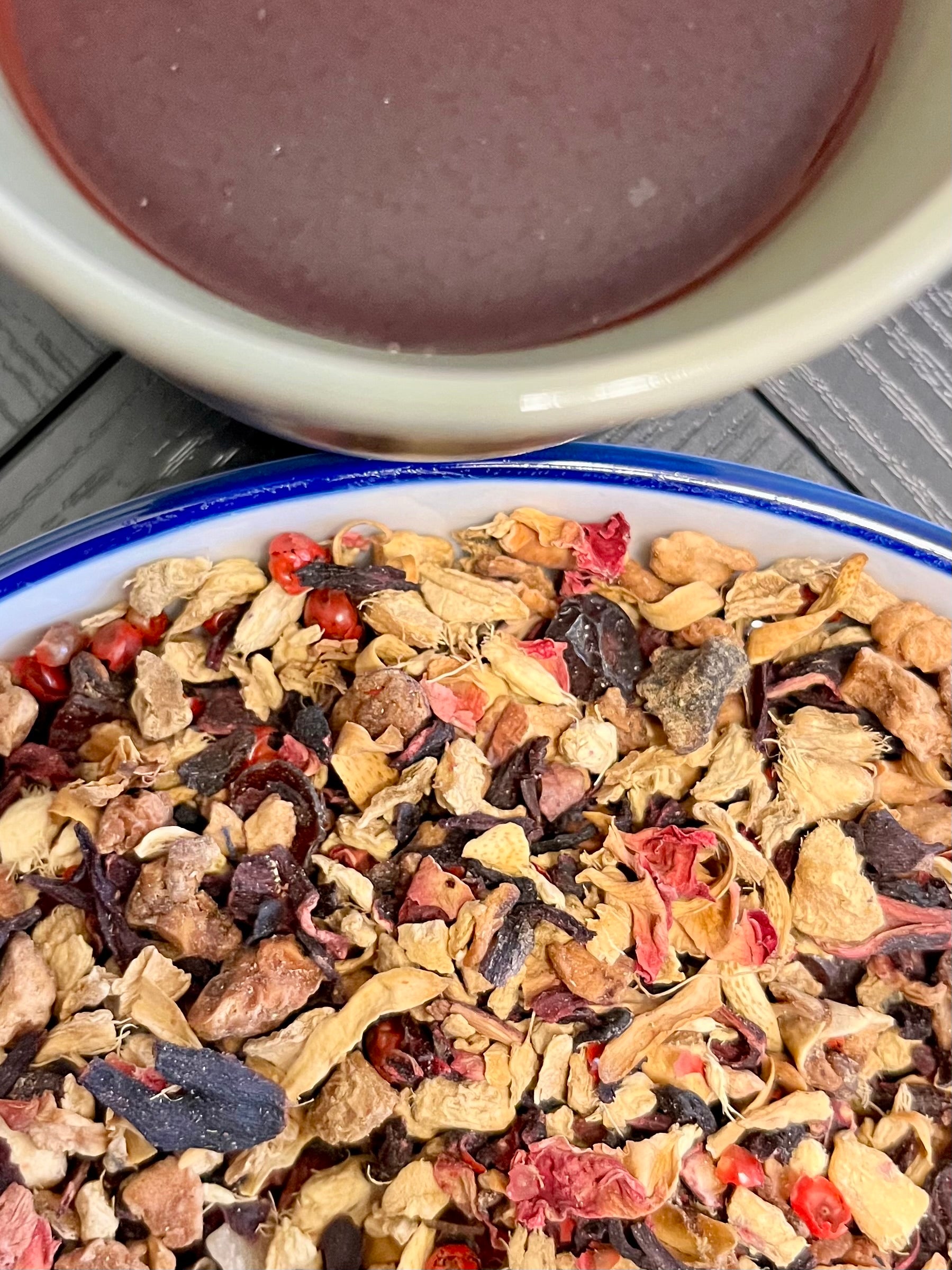 Tea leaves with visible herbs and fruits in a bowl, next to a cup of tea.